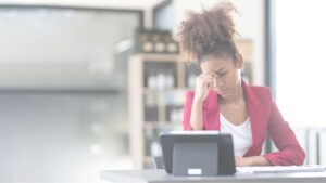 A imagem mostra uma mulher jovem sentada em uma mesa, olhando para um tablet com uma expressão de preocupação. Ela usa um blazer vermelho sobre uma camiseta branca e está com uma das mãos apoiada no rosto, tocando a testa. O ambiente ao fundo parece ser um escritório moderno, com uma estante desfocada, que sugere um ambiente de trabalho profissional. A iluminação é suave, com tons claros predominando na composição, que destaca o contraste entre o blazer vermelho e o cenário mais neutro.