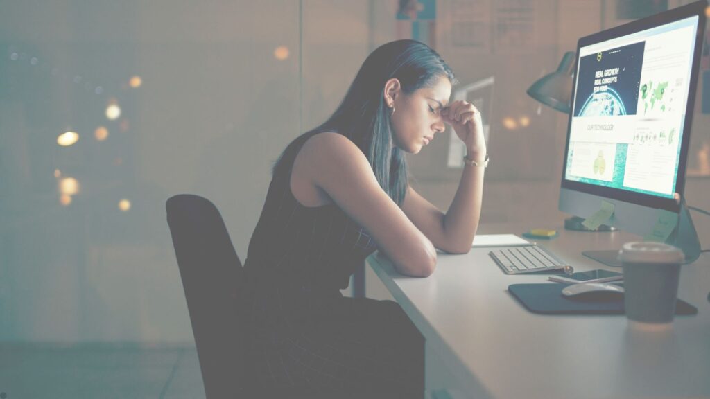 A imagem mostra uma mulher sentada em frente a um computador, com uma expressão de cansaço e estresse. Ela apoia a cabeça com uma das mãos e parece pensativa ou exausta. O ambiente parece ser um escritório, com luzes ao fundo indicando que é noite ou um horário com baixa iluminação externa. Na mesa, há um copo descartável, teclado, mouse, celular e alguns papéis ou post-its.