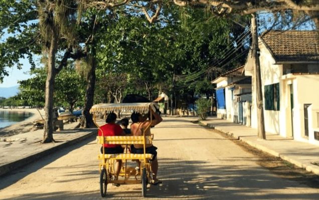 imagem de uma pequena charrete puxada por bicicleta em uma estradinha de chão cercada de árvores. Do lado esquerdo é possível ver o mar e do lado direito algumas casas. Foto foi tirada na Ilha de Paquetá cidade do Rio de Janeiro